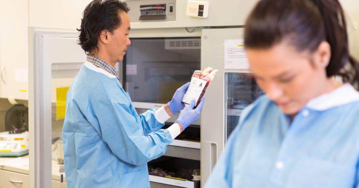 The Importance of Temperature Monitoring for Blood Banks A healthcare worker wearing gloves to examine a labeled blood bag in refrigerated storage while another worker stands nearby.