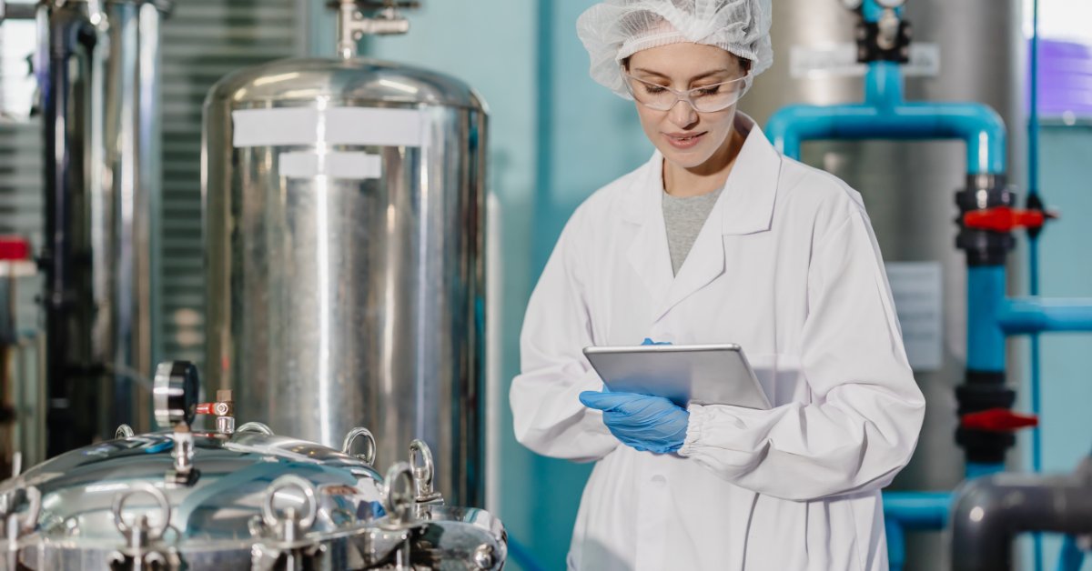 A laboratory worker in a white coat, hair net, safety glasses, and gloves using a tablet beside metal tanks.