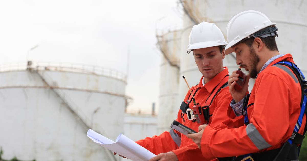 In front of several oil refinery storage tanks, two engineers wearing orange protection clothing examine a blueprint.