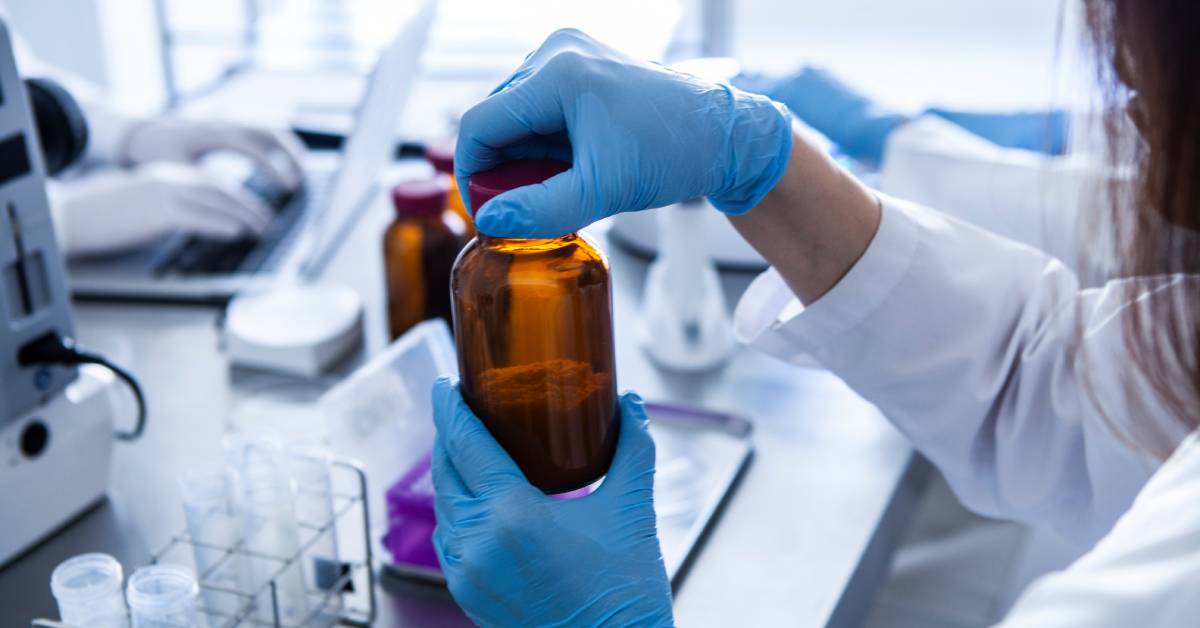 In a laboratory, a woman wearing sterile blue gloves and a lab coat opens the lid of an amber bottle filled with liquid.