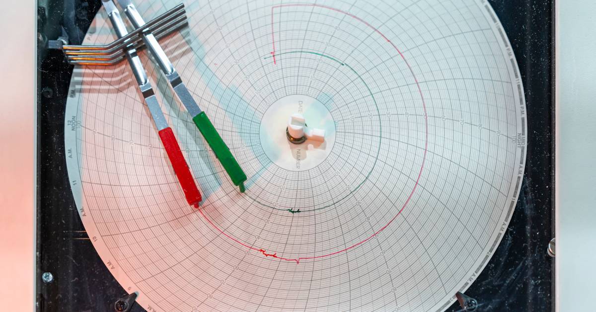 A green and red pen records temperature data on a sheet of circular chart recorder paper against a black background.