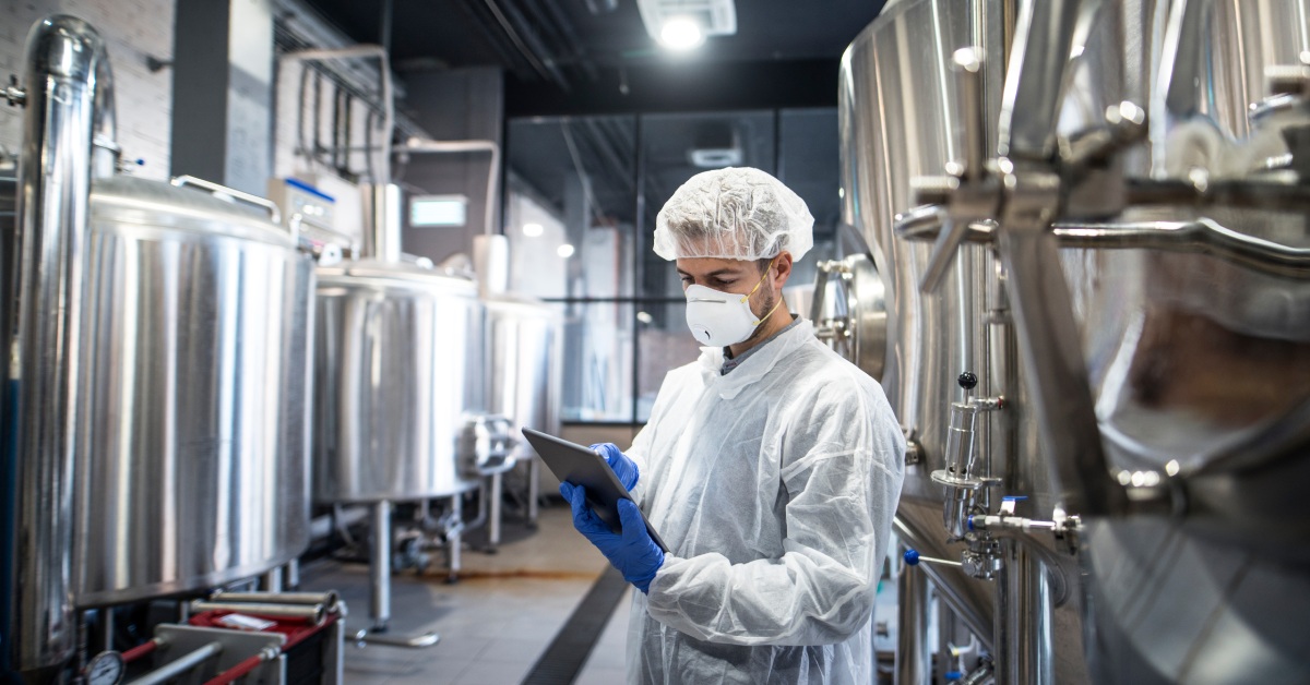 A pharmacist is surrounded by large metal containers. He wears white protective clothing and uses a tablet with blue gloves.