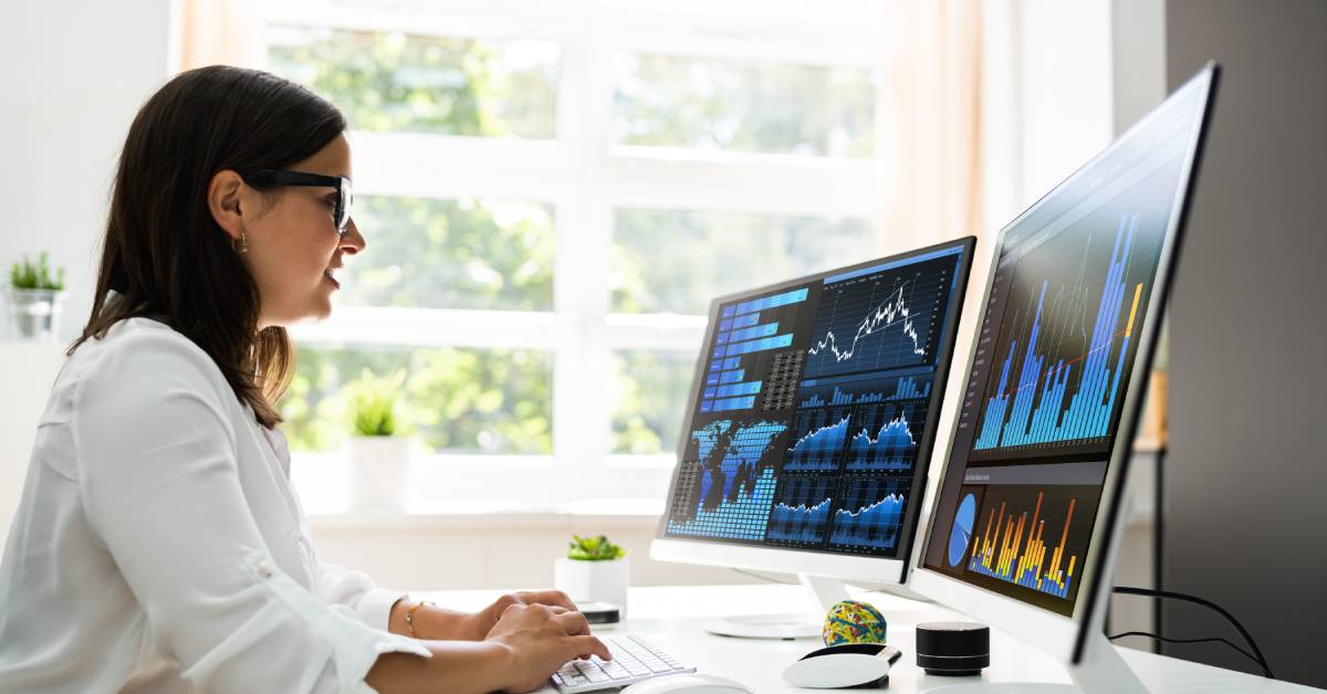 A woman wearing glasses analyzing data while sitting at her desk. There are multiple graphs and charts on her computer.