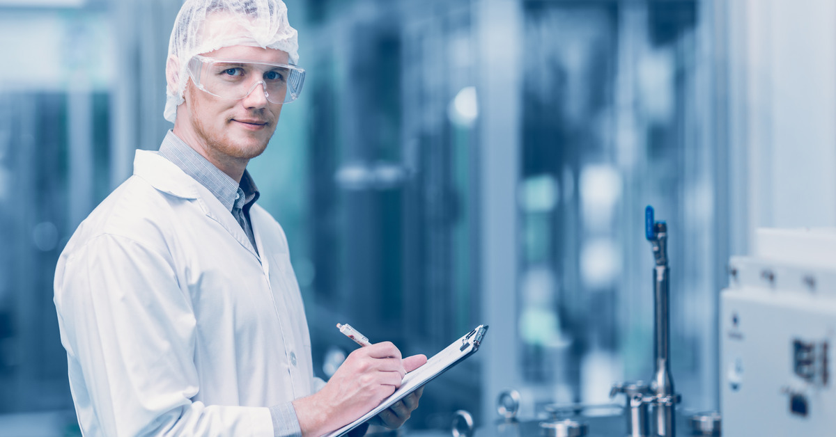 A laboratory worker wearing a white coat, a hairnet, and goggles, staring into the camera while writing on their clipboard.