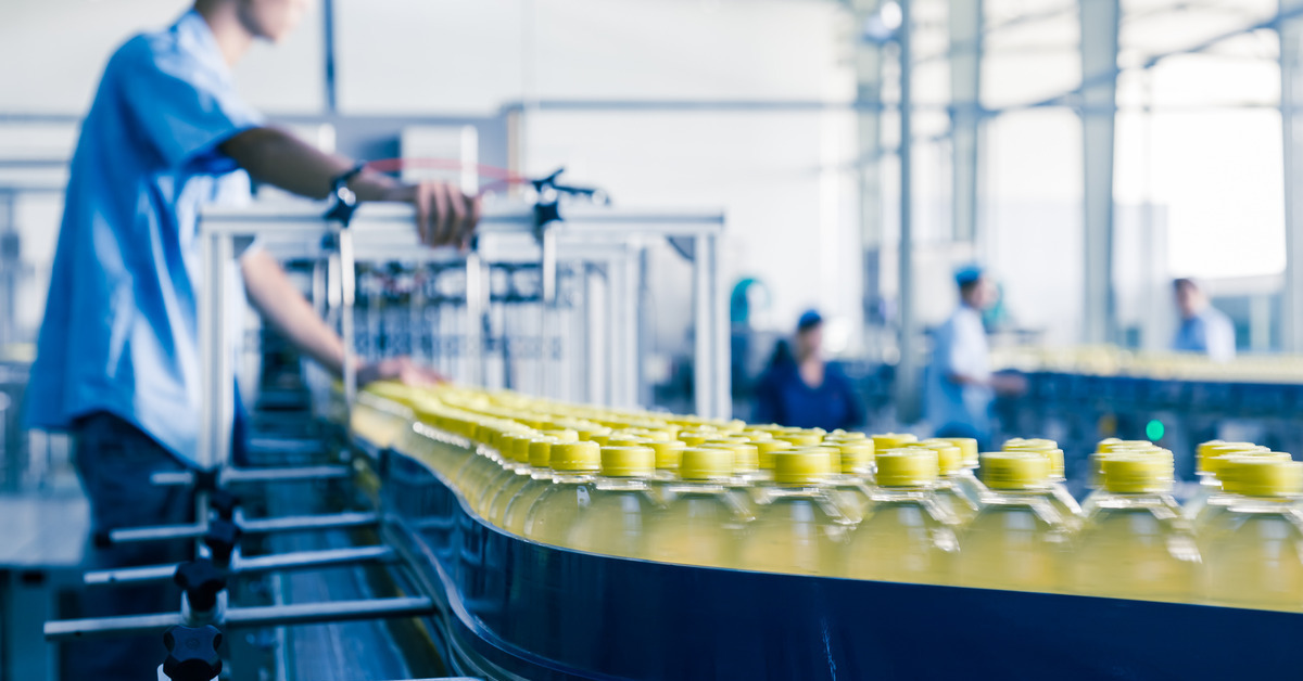 Many bottles travel down an assembly line in a manufacturing plant. A worker wearing a blue shirt oversees the process.