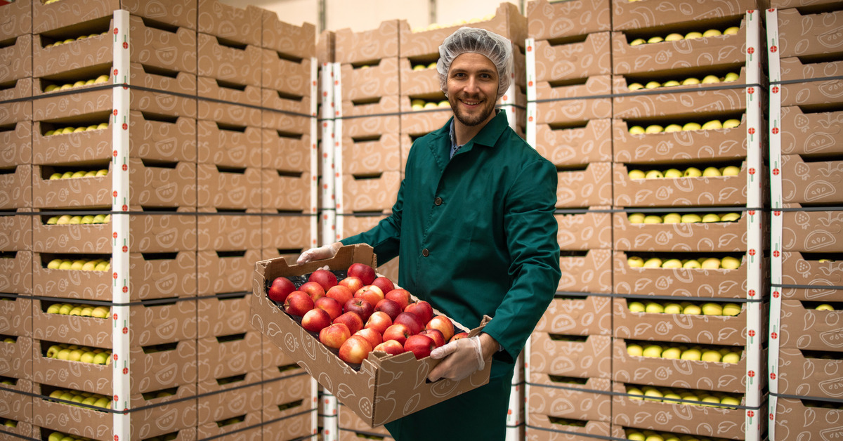 A worker wearing a green jacket, hairnet, and gloves holds a crate of apples. Many stacked apple crates appear in the background.