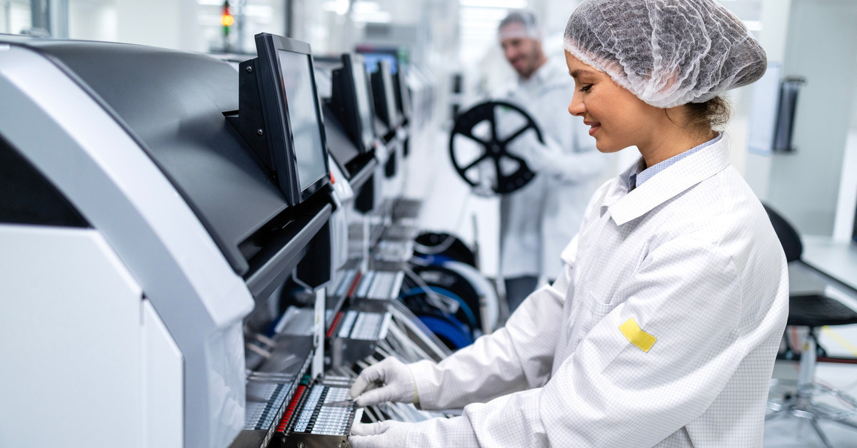 A woman wearing a white hairnet and coat operating a semiconductor production machine. A man works in the background.