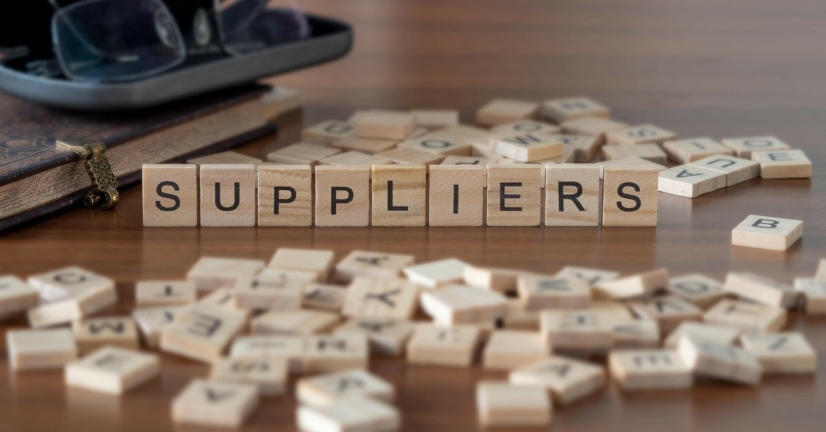 Wood letter tiles scattered on a wooden table. Some of the letter tiles are standing upright to spell the word 'Suppliers.'