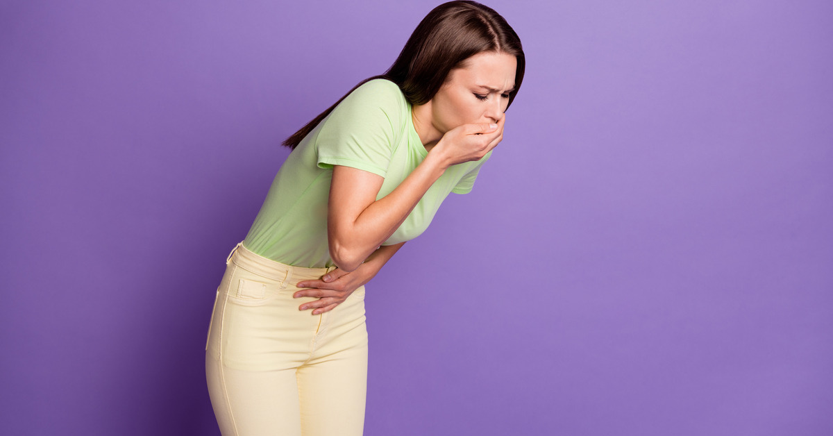 A dark-haired woman in a green shirt hunches over. Her right-hand covers her mouth, and her left hand is on her stomach.