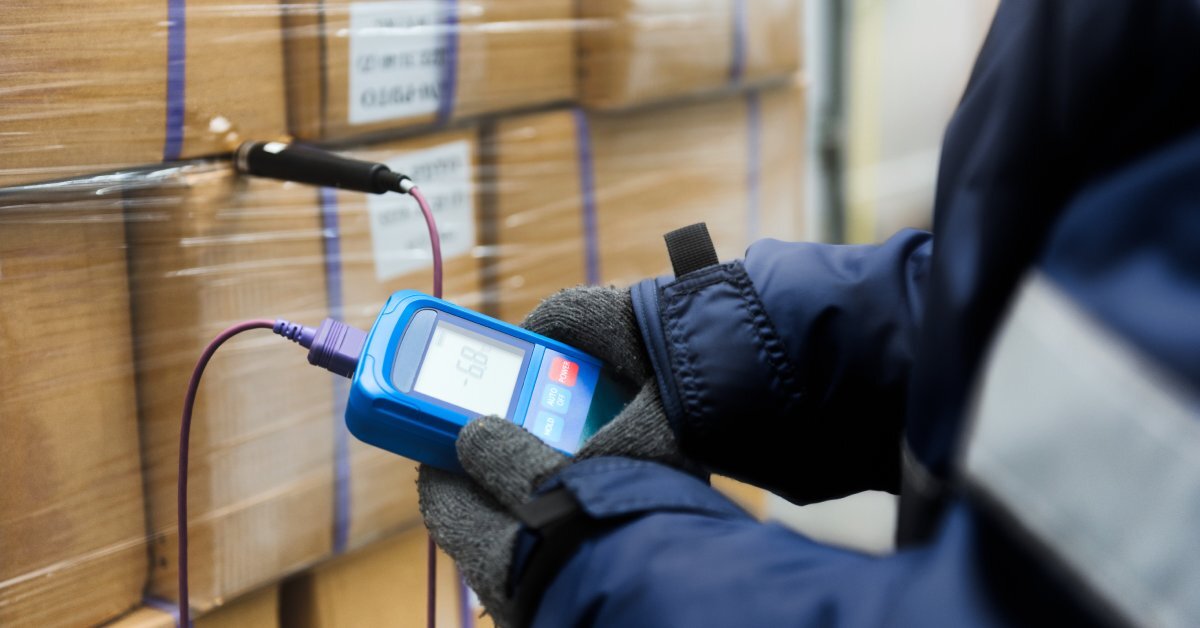 A worker wearing a jacket and gloves uses a thermometer to measure the temperature of boxes containing food products.