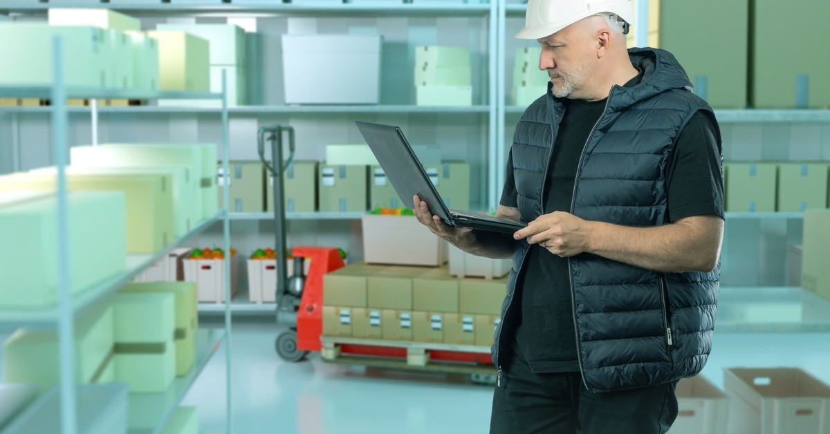 A man in a white hard hat and black vest holding an open laptop while standing in a large cold storage room. A man in a white hard hat and black vest holding an open laptop while standing in a large cold storage room.