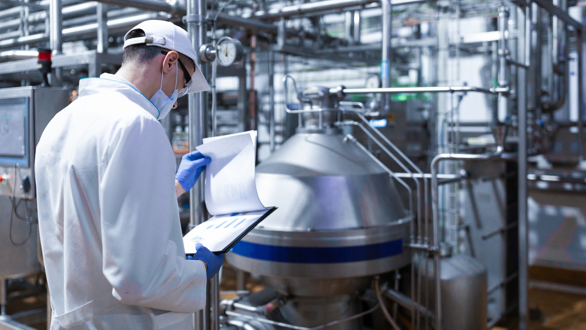 A worker in a white lab coat, white hat, blue gloves, and face mask looking at a clipboard in front of a large silver machine.