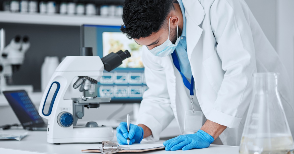 A man in a white lab coat, medical mask, and blue gloves standing by a microscope while writing on a clipboard.