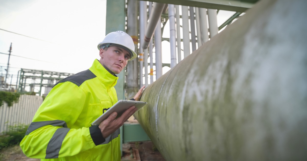 A worker wearing a hard hat and safety jacket while holding a tablet and inspecting a large pipeline outside.