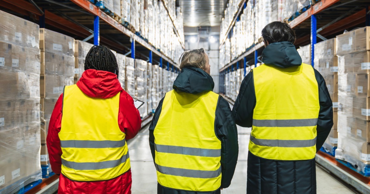 Three people wearing high-visibility vests over long jackets while standing together in a cold storage warehouse.