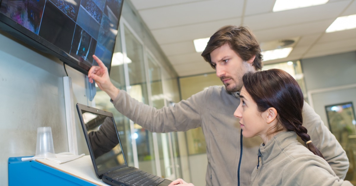 Two people standing in front of a laptop and a screen showing multiple locations. Each person is looking at a different screen.