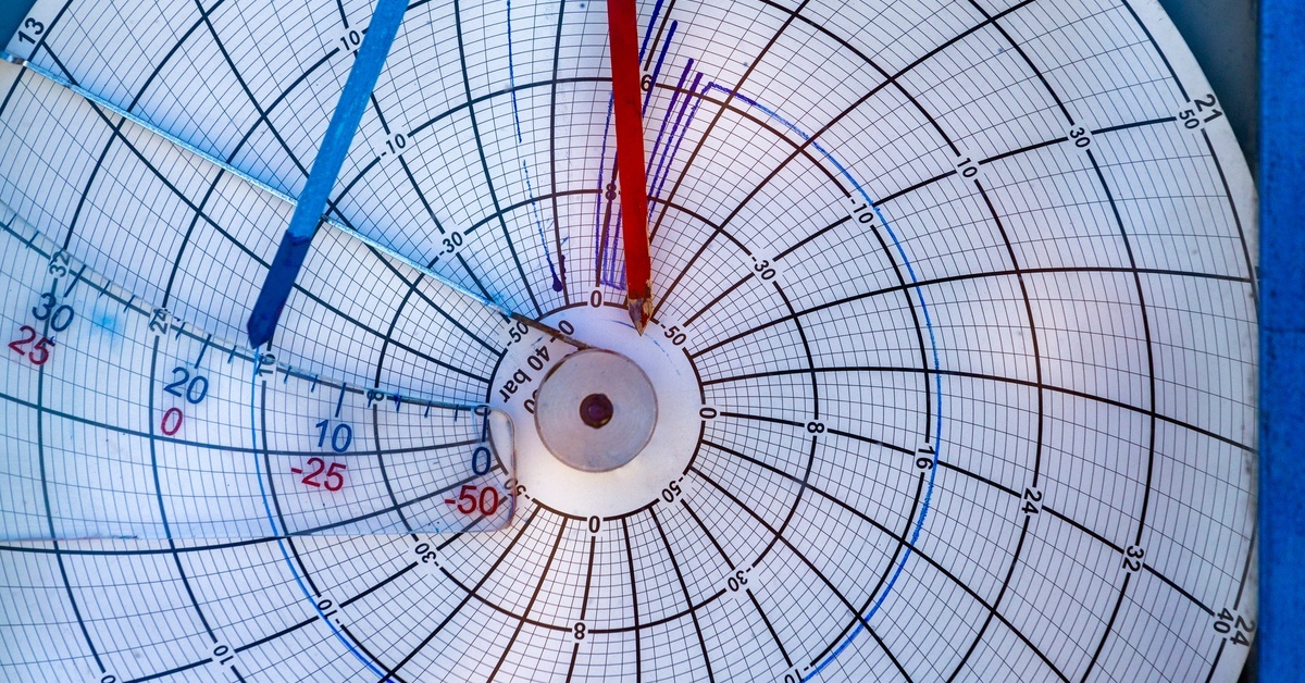 A circular chart recorder, up close, with one red and one blue pointer and chart paper, capturing pressure readings.